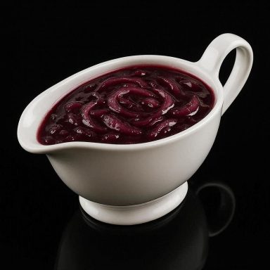 Rotwein‑Zwiebel‑Sauce mit sichtbaren Zwiebelstückchen in einer weißen Porzellan‑Sauciere auf schwarzem, spiegelndem Untergrund.