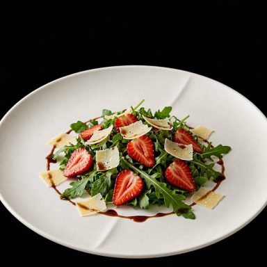 Erdbeer-Rucola-Salat Erdbeer-Rucola-Salat mit gehobeltem Grana Padano und feiner Balsamico-Reduktion, frisch angerichtet auf einem Teller.