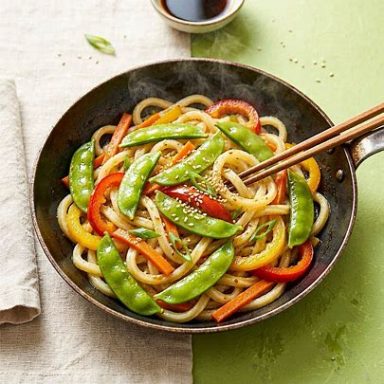 🥢 Asiatische Udon‑Gemüsepfanne Bunte Udon‑Gemüsepfanne mit Zuckerschoten, Karotten, Paprika und Sesam in einer Pfanne serviert, glänzend, frisch und leicht gebraten.